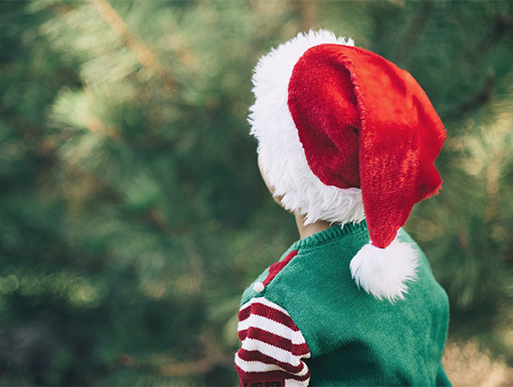 Child in a green festive jumper and red santa hat looking at an uncut christmas tree
