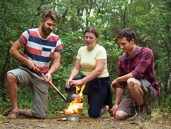 Two men and a woman in a luscious forest working together to build a fire and make tea.