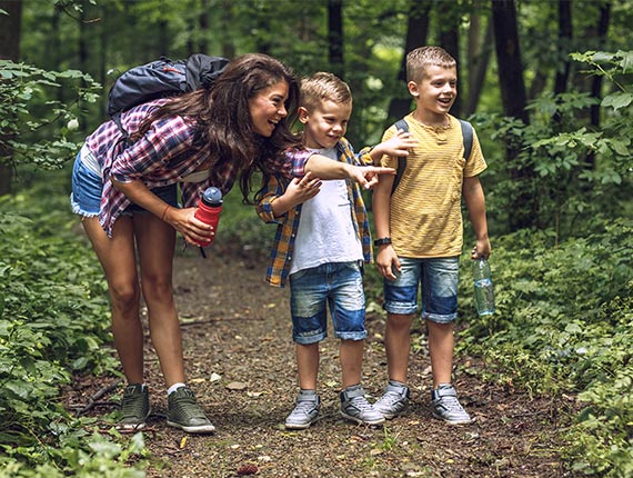 An adult and two children on a woodland path with water bottles and backpacks. The adult is pointing to something off-camera and the children are looking over in the same direction.