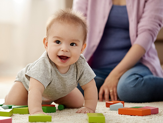 Smiling baby crawling amongst toys as his mother watches on.