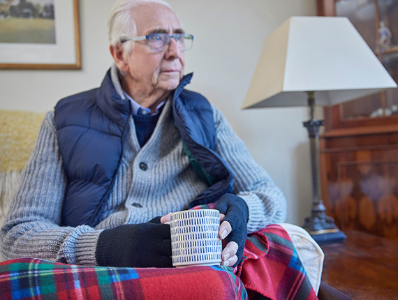 Older man sat in his living room wrapped up in a blanket and wam jumpers wearing gloves and holding a hot cup of tea.