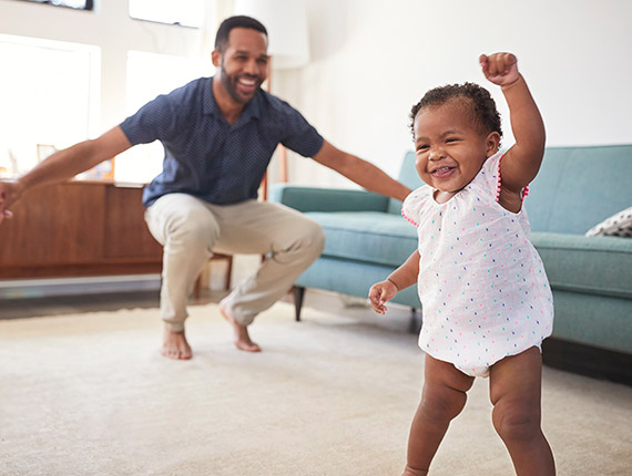Baby walking across living room with a smile and dad in the background looking happy.