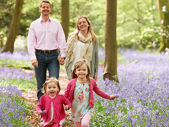 Mum, dad and 2 young daughters smiling as they walk through a woodland filled with bluebells.